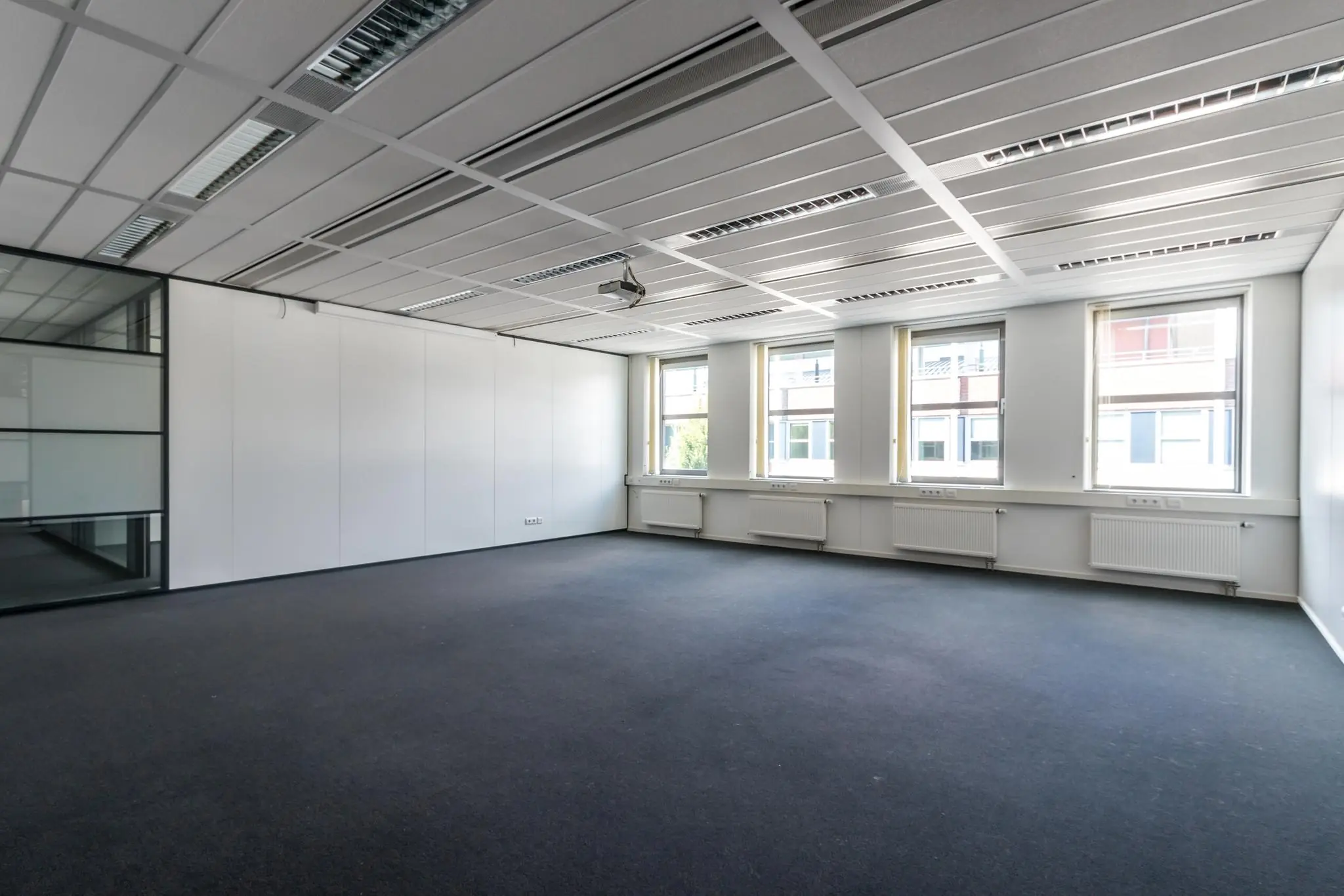 Lichte, lege kantoorruimte met drie grote ramen, witgesausde muren en systeemplafond met ingebouwde verlichting.